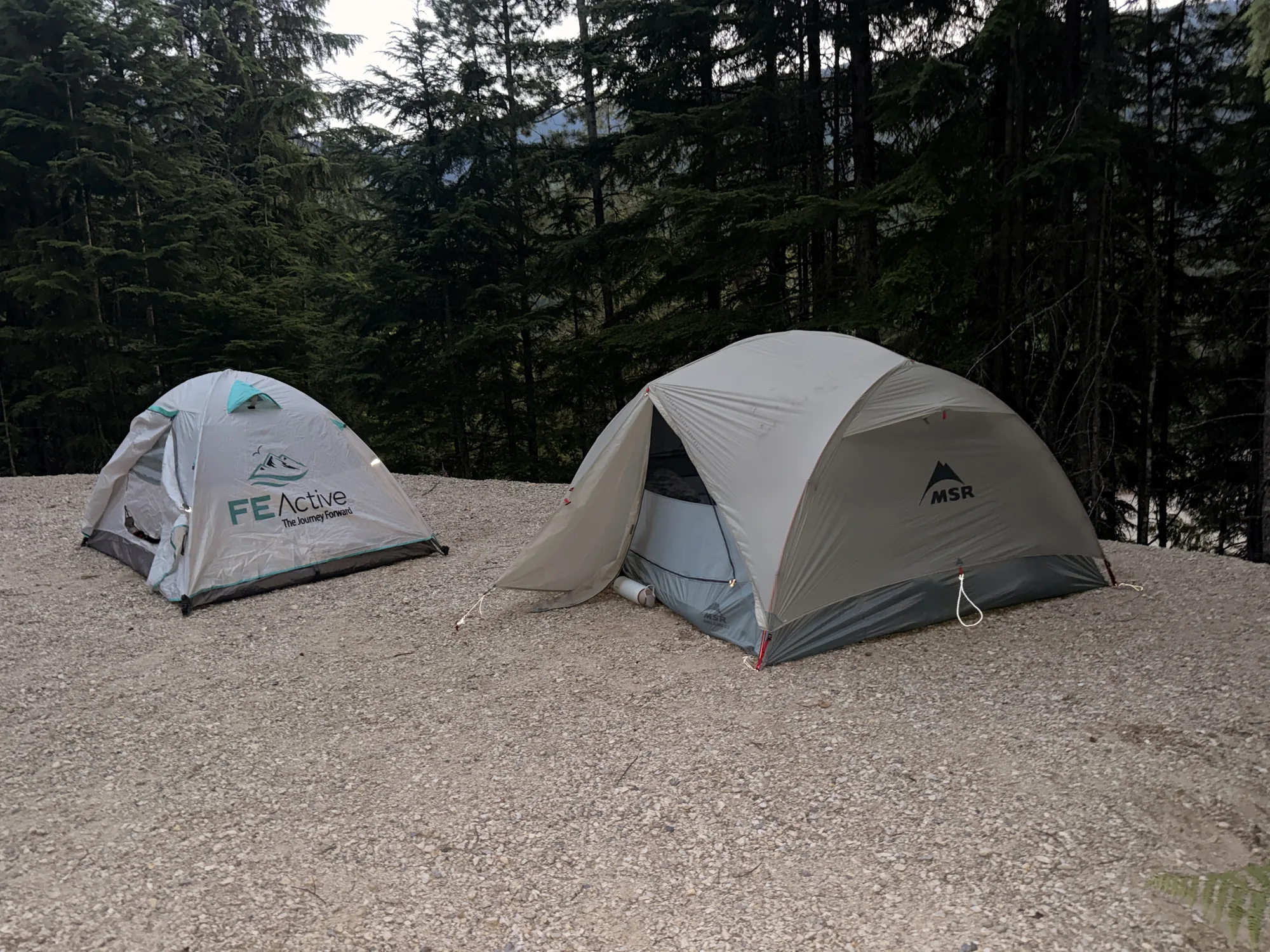 Two tents pitched on a gravel campsite pad surrounded by forest